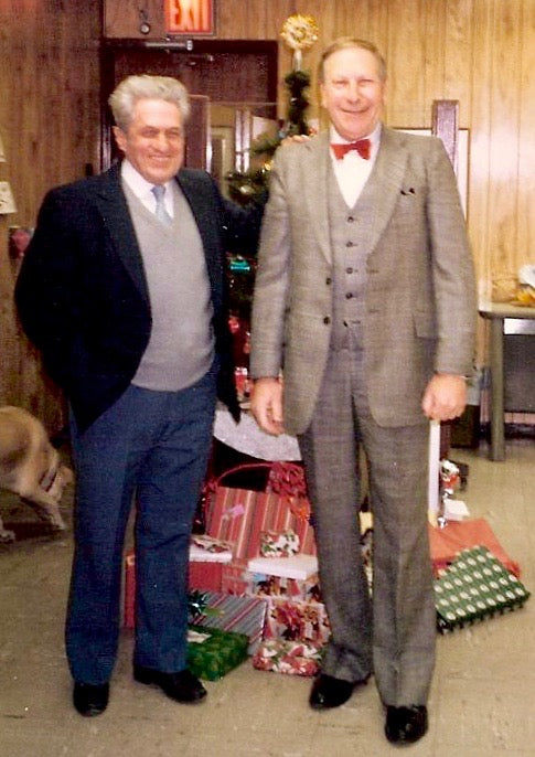 Abe and Fred in suits standing in a room with Christmas decorations and presents.